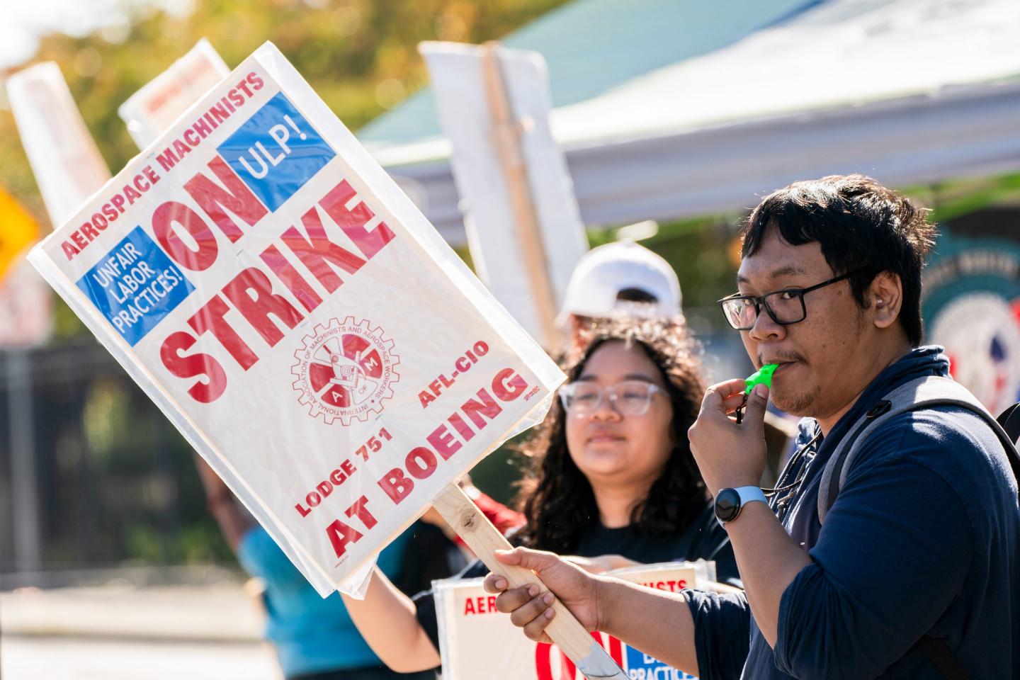 A man blows a whistle while holding a sign as he and other workers picket during a strike.