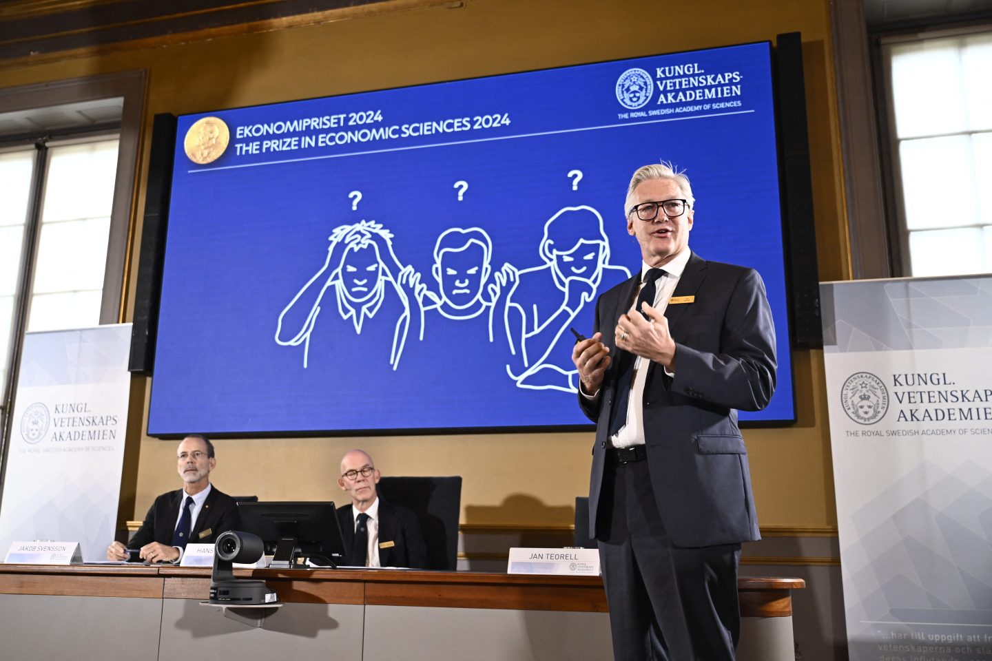 Academy of Sciences permanent secretary Jan Teorel delivers a press conference in front of a screen showing results of the research of laureates of the 2024 Sveriges Riksbank Prize in Economic Sciences in Memory of Alfred Nobel, Daron Acemoglu, Simon Johnson and James A Robinson