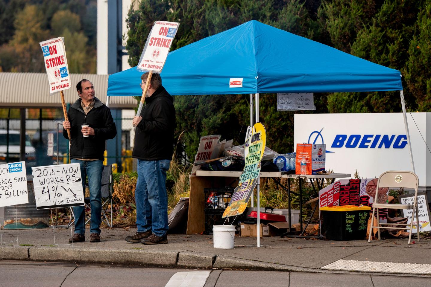 Two men talk and hold picket signs at a table and tent set outside Boeing's manufacturing facility.