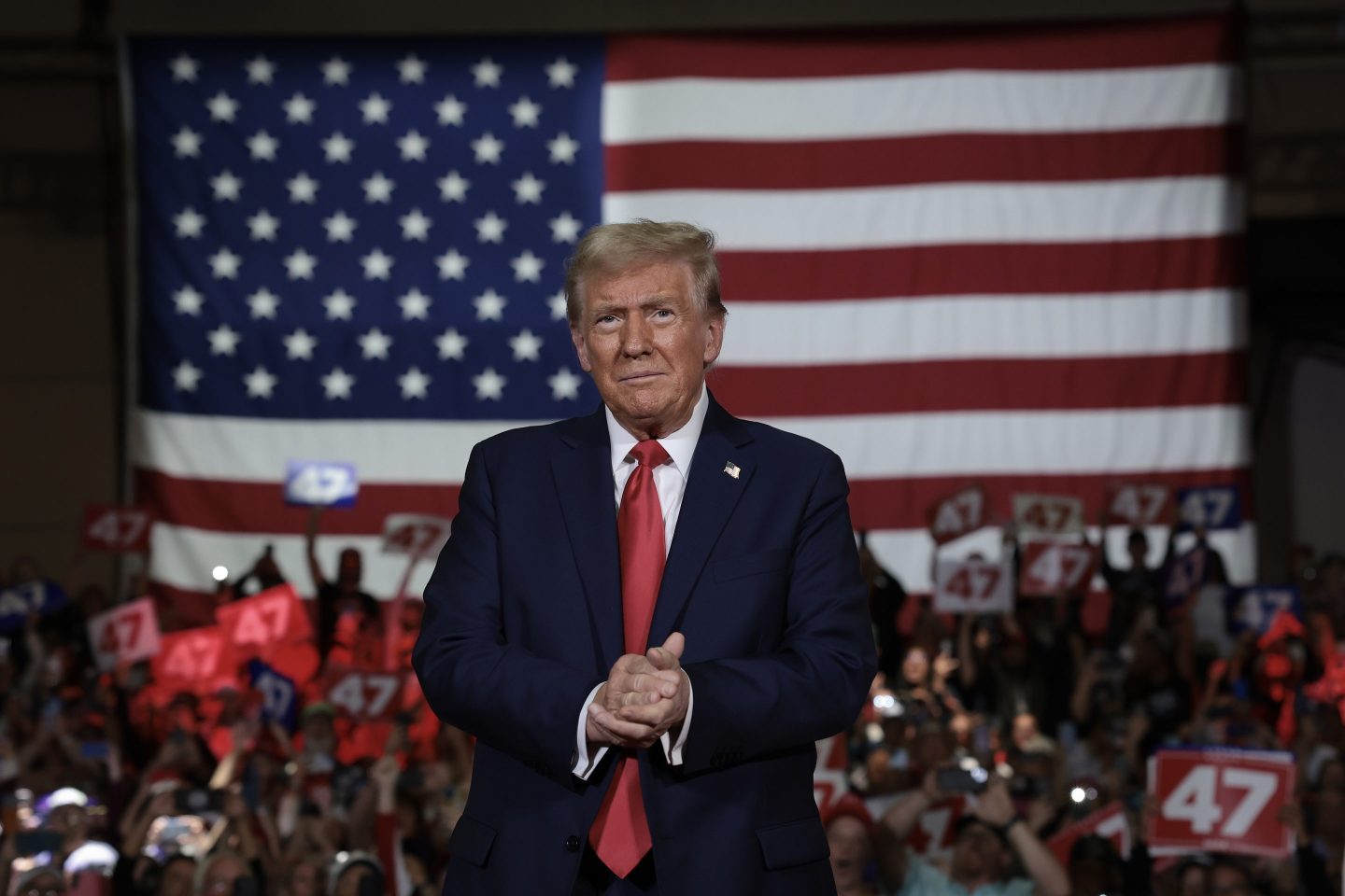 Donald Trump clasps his hands together standing in front of a crowd and large American flag.