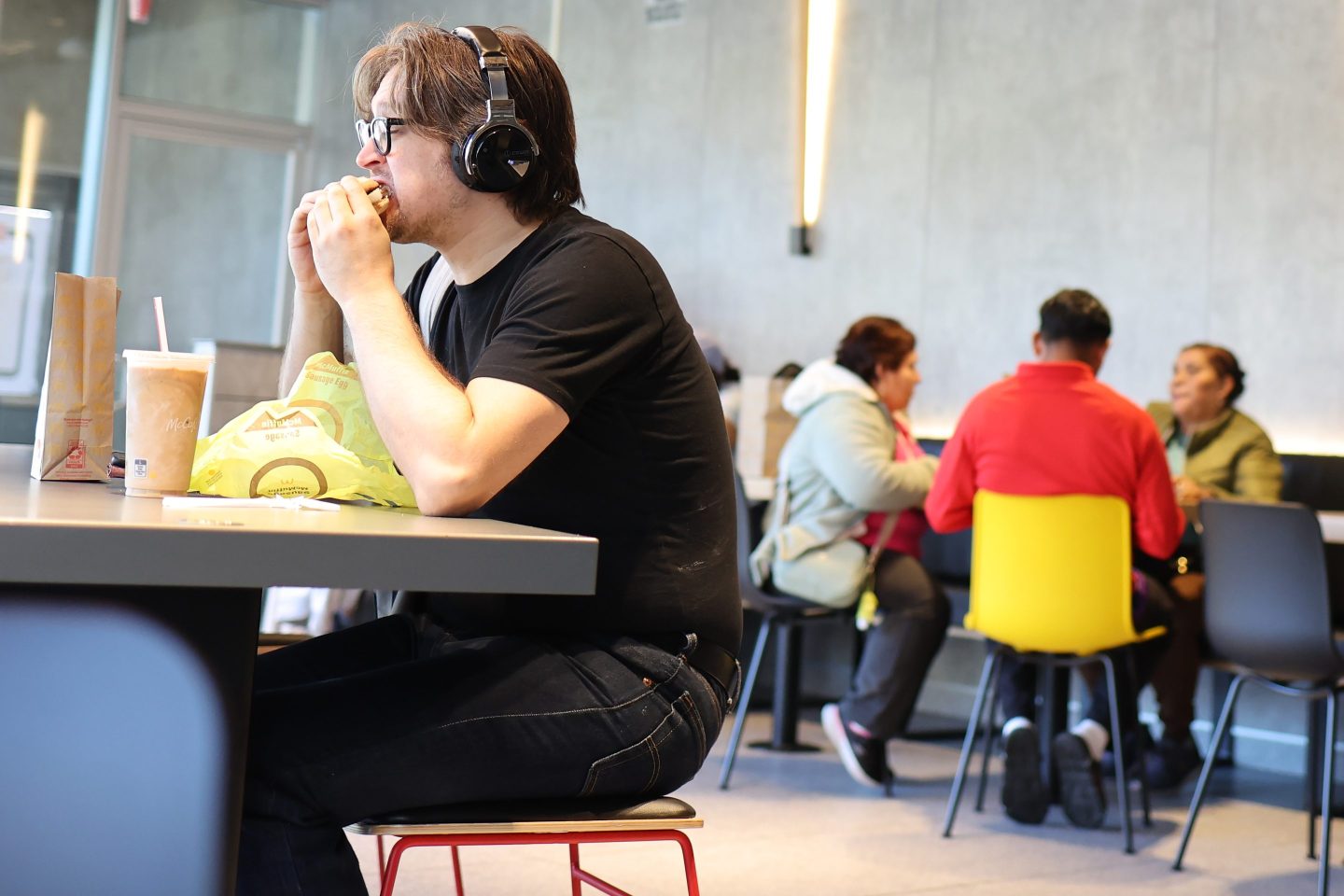 A man wearing headphones is sitting at a table eating a burger.
