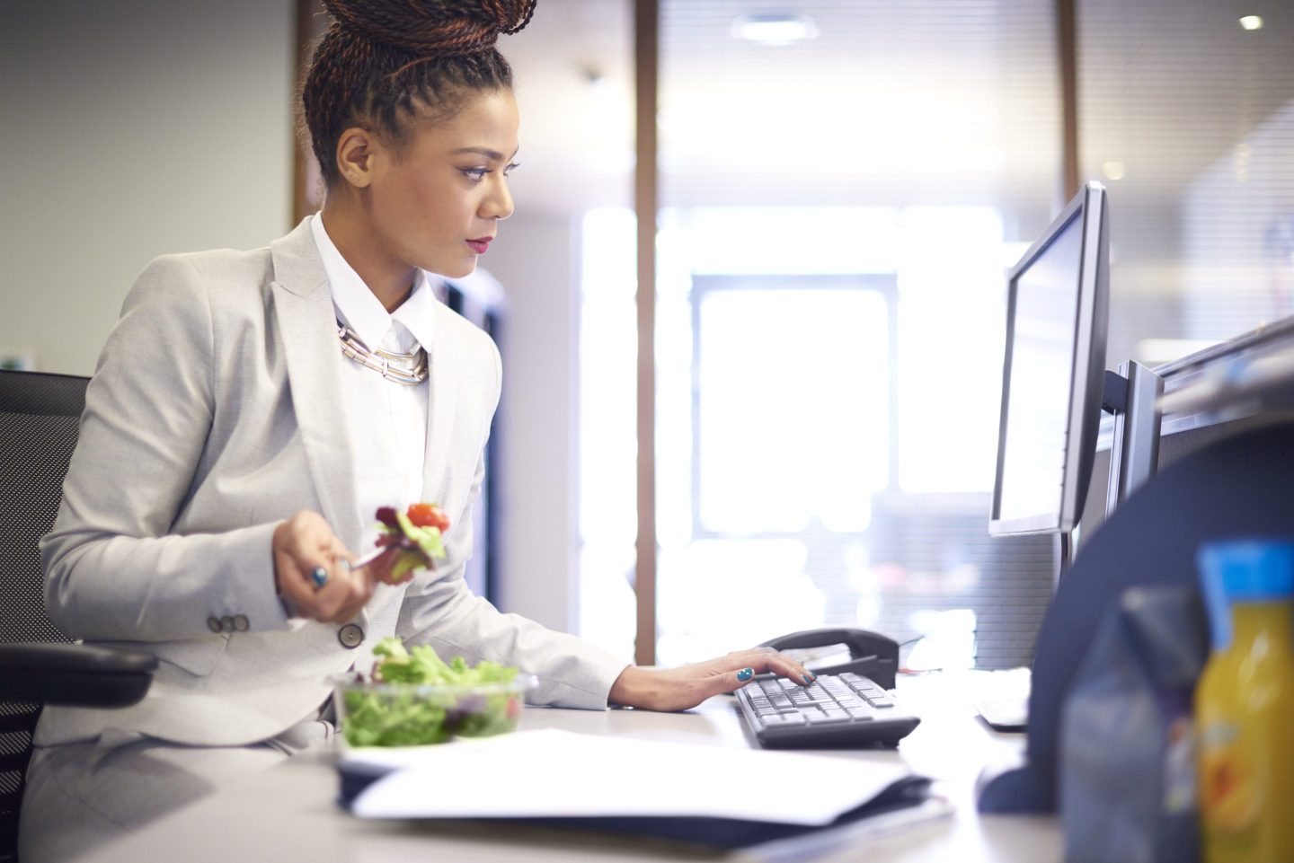 A businesswoman sits at her desk checking her computer whilst eating a salad from a salad.