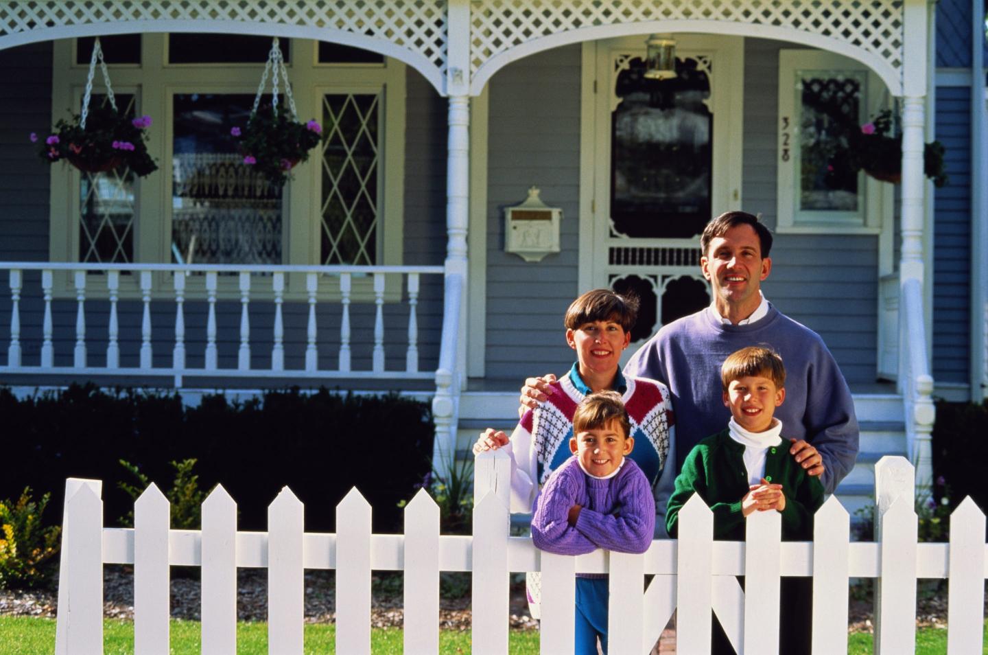 A family stand outside their home, leaning on a white picket fence.
