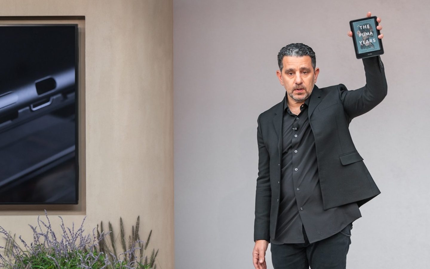 Amazon executive Panos Panay holds up the new Kindle Colorsoft device on stage, with a TV screen displaying video of the device behind him