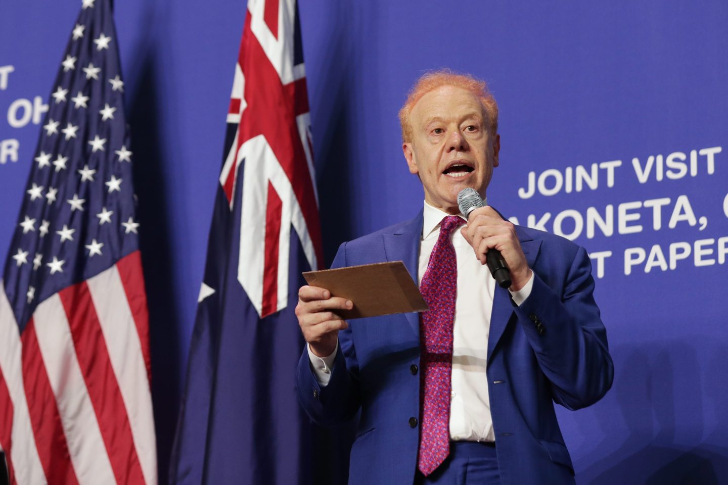 Anthony Pratt stands in front of American and Australian flags and reads off a card into a mic.