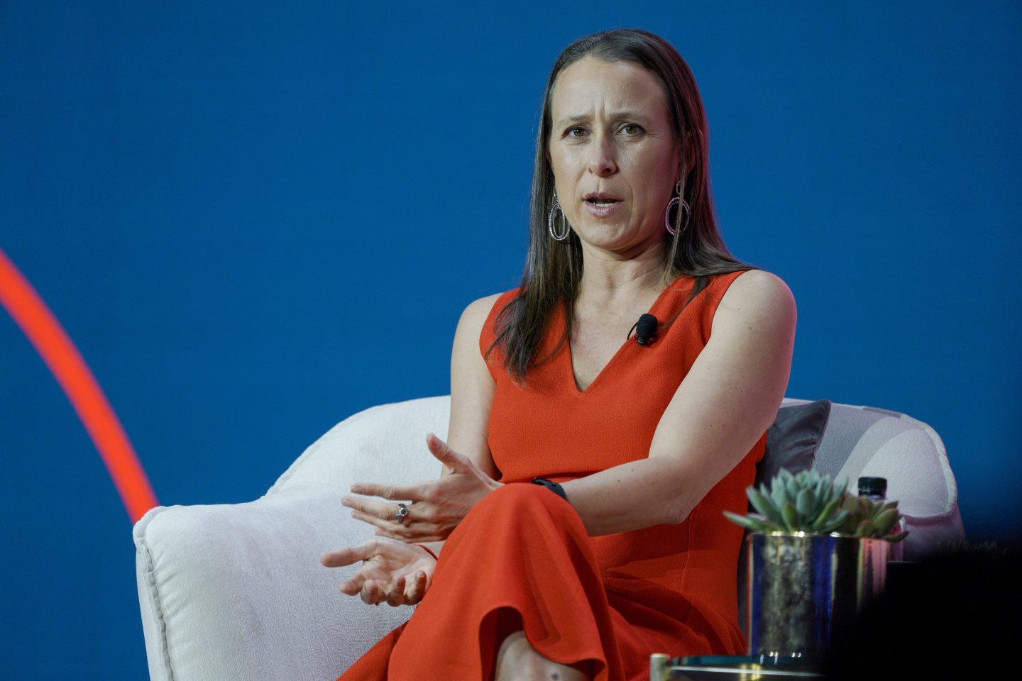 Anne Wojcicki, wearing a red dress, sits in a white chair as she speaks.