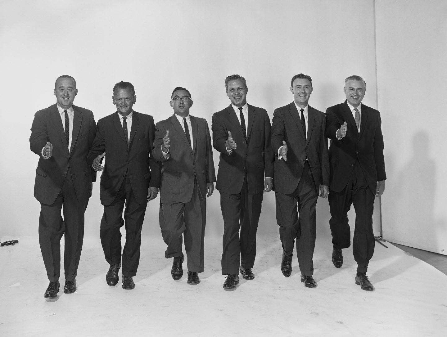 Row of six businessmen extending hand to shake, smiling.