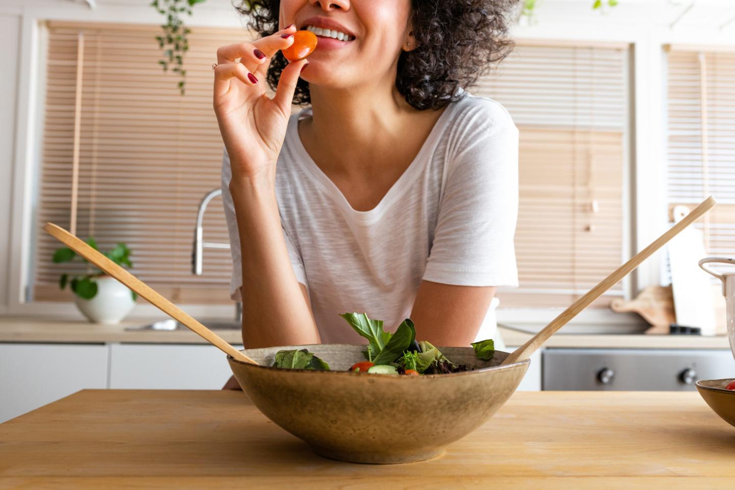 woman leaning on a counter eating a tomato out of a salad