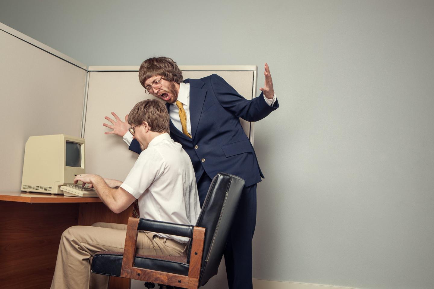 A manager stands next to his employee's desk and yells angrily at him.