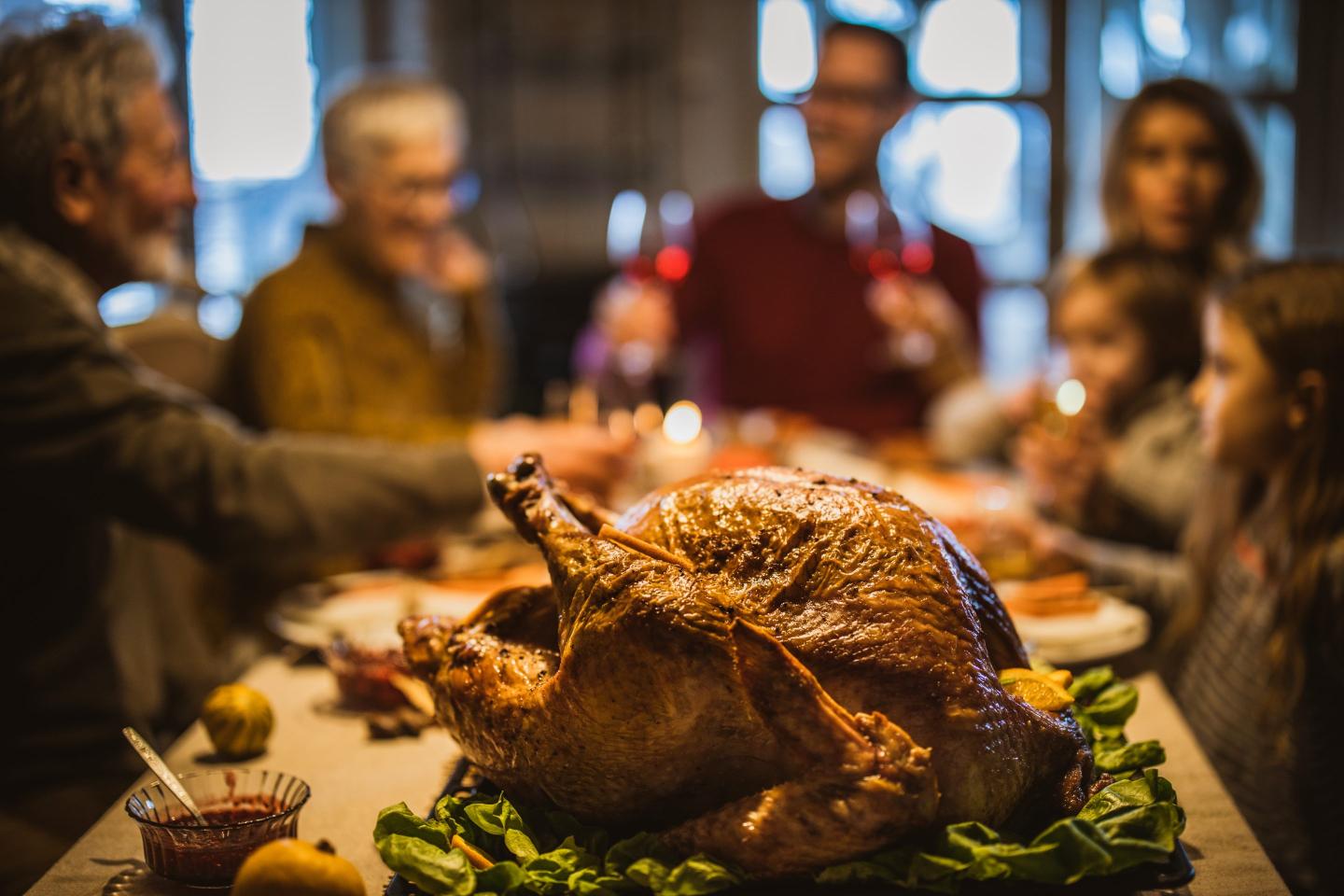 Thankgiving turkey is seen in the foreground as a family in the background celebrates.