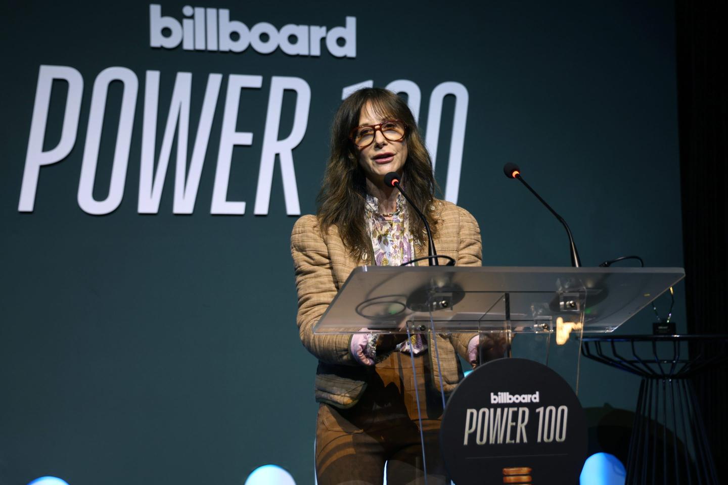 A woman with brown hair stands behind a clear lectern with a plaque that reads "Billboard Power 100"