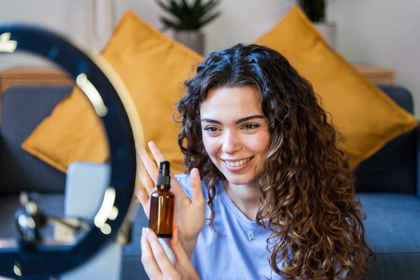 A woman holds up a skincare product in front of a ring light and camera.