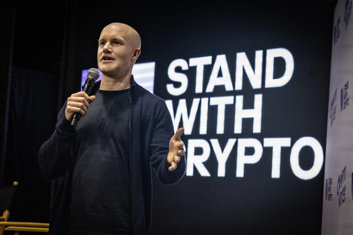 businessman speaks onstage with a "stand with crypto" screen behind him