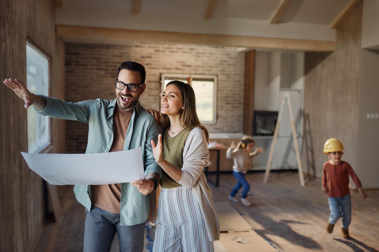 Happy couple cooperating while reading blueprints and making plans in their renovating house. Their kids are in the background.