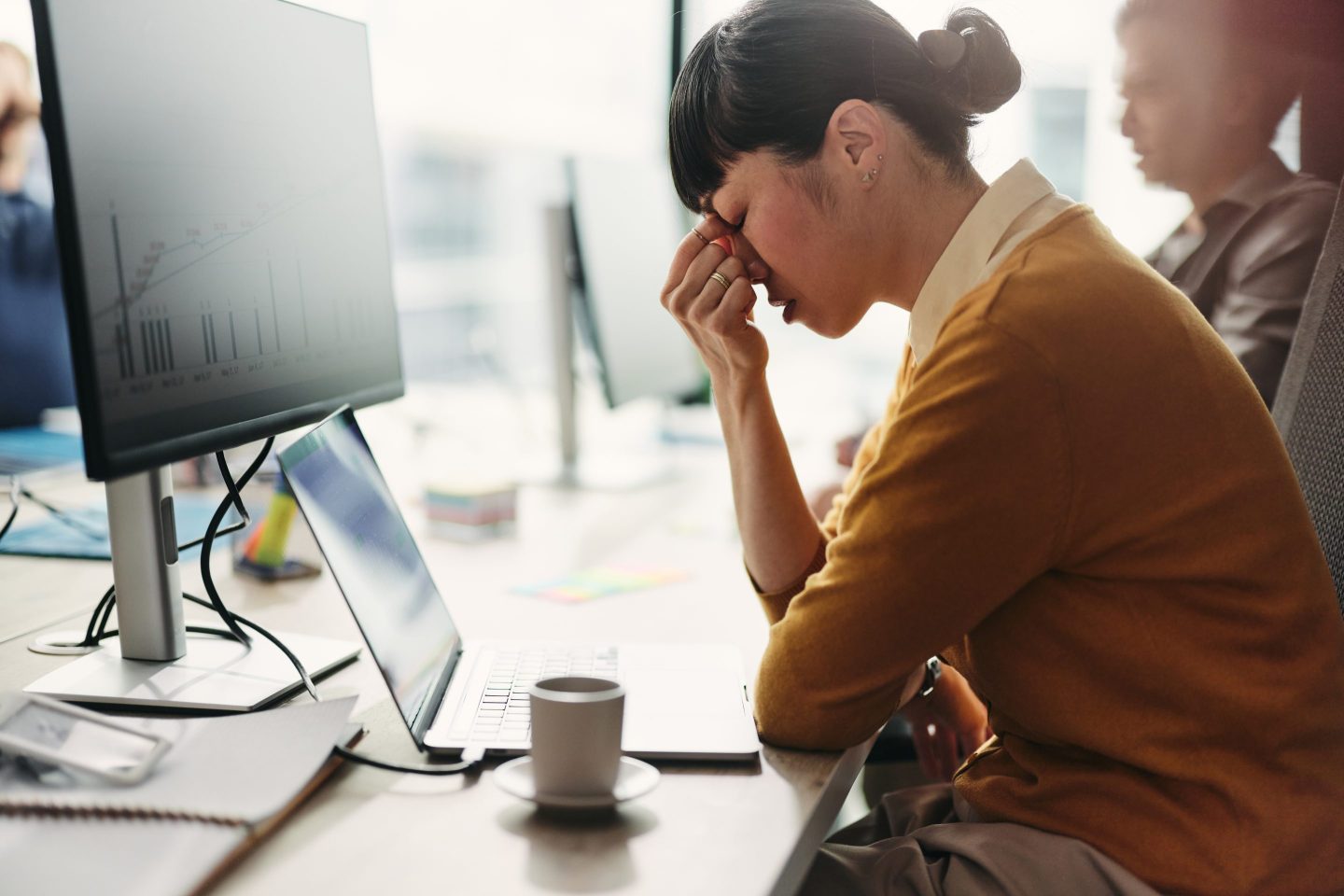 Exhausted Asian female programmer holding her head in pain while working desktop PC at corporate office.
