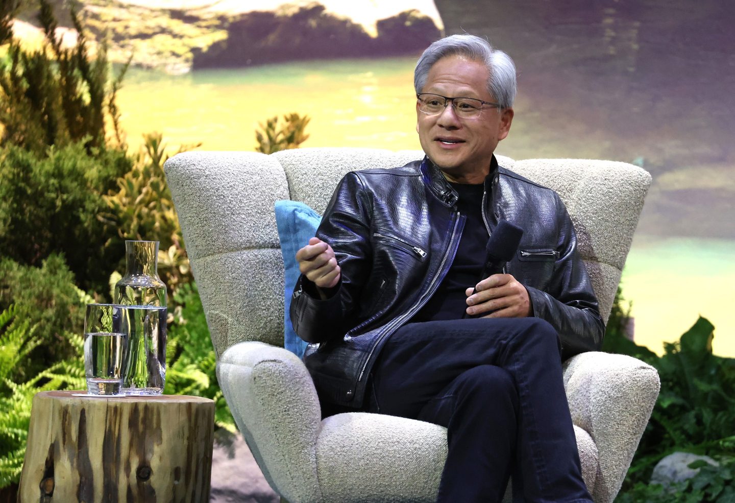 Jensen Huang talks onstage in a big chair with greenery and a projection of nature in the background.