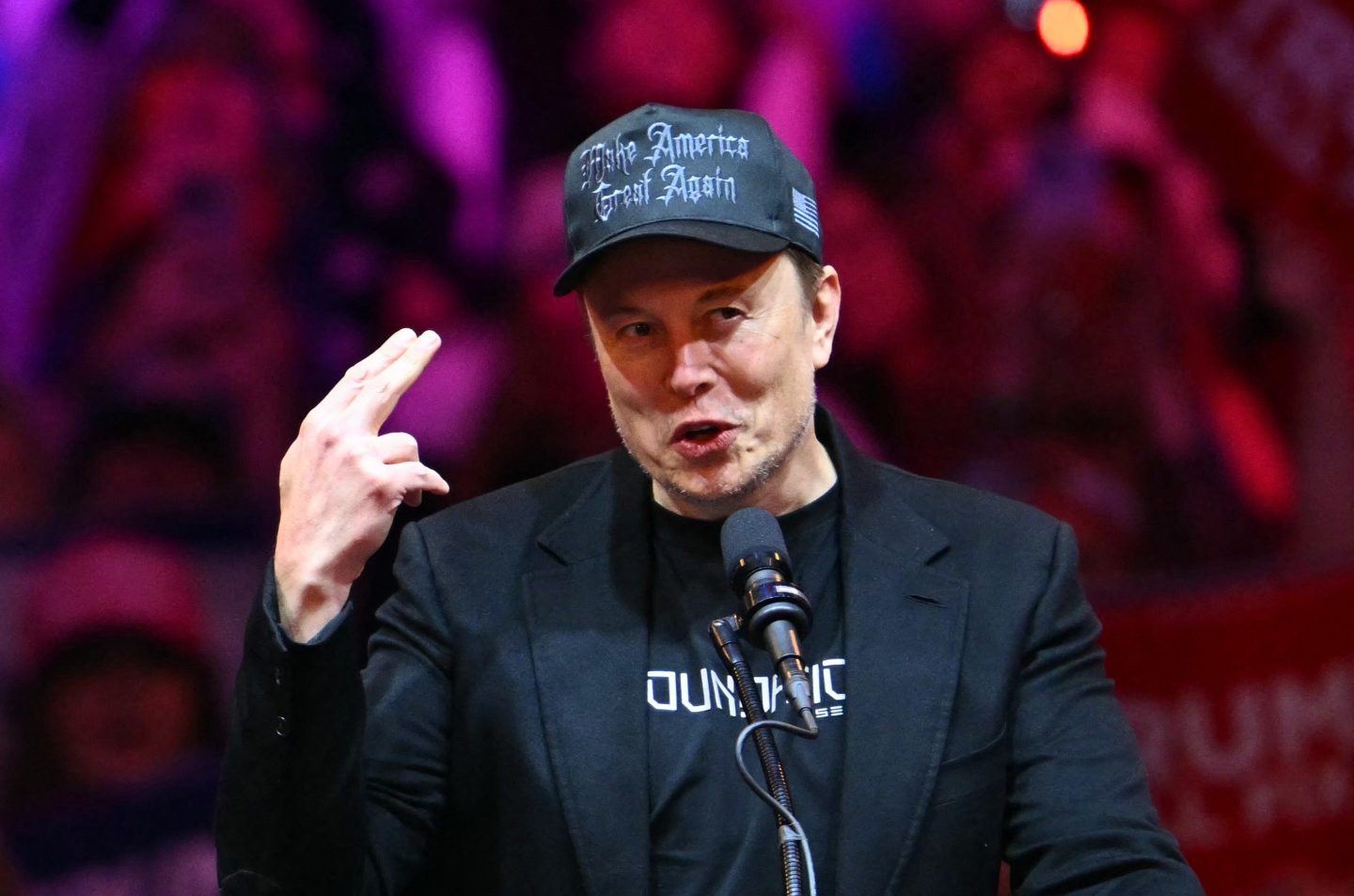 Elon Musk in a black "Make American Great Again" hat pointing two fingers in the air.