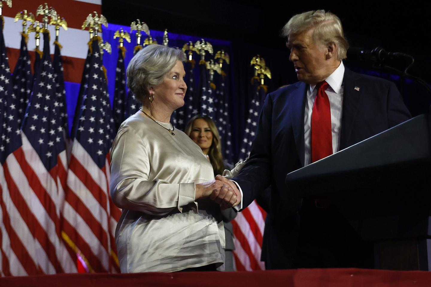 Susie Wiles, Trump's campaign manger, shakes his hand on stage near a podium.