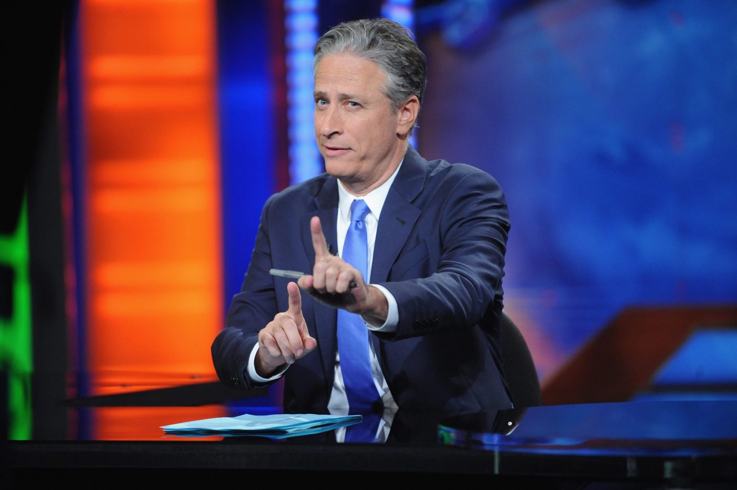 Jon Stewart sits behind a desk and holds his hands up, pointing.