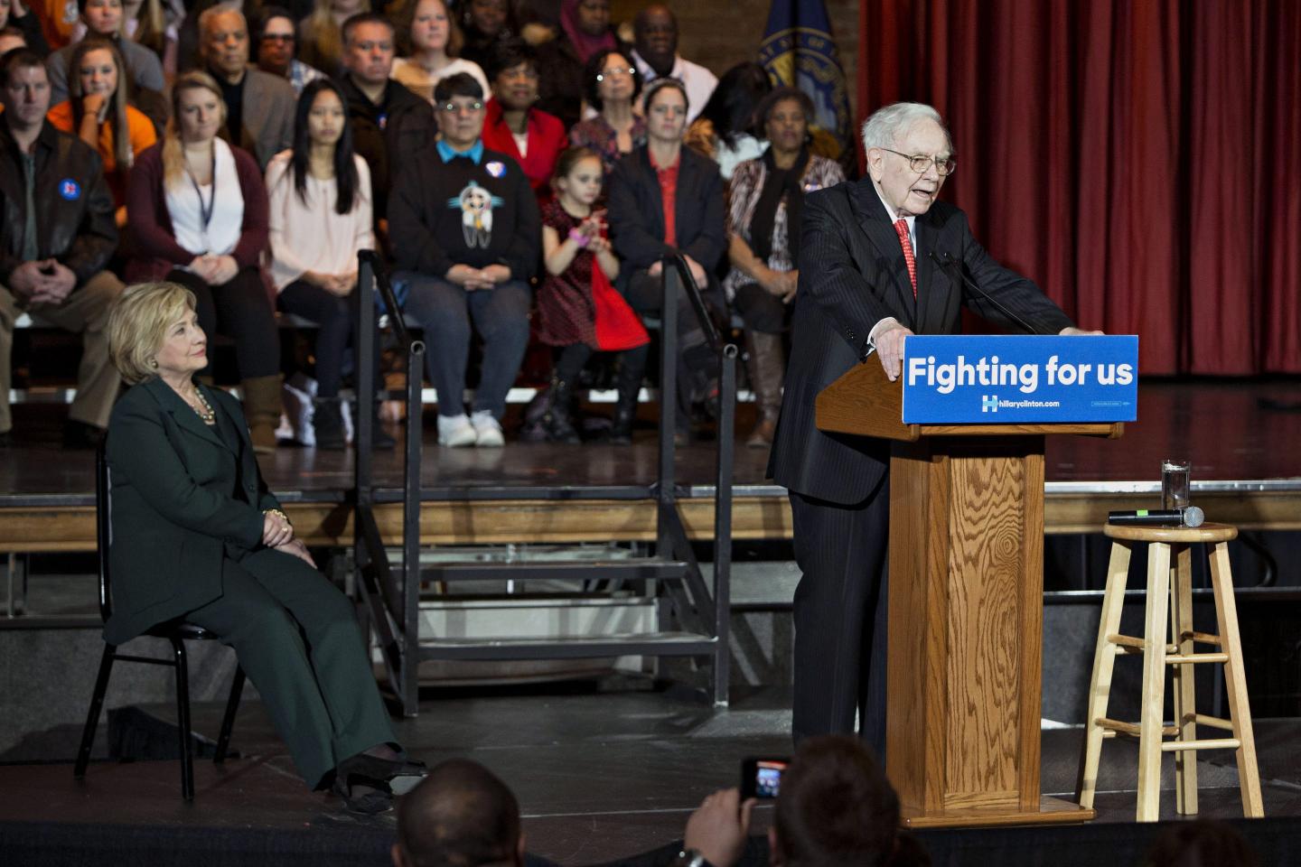 Warren Buffett speaks at a podium with a crowd both behind and in front of him as Hillary Clinton watches on from a chair to his left.