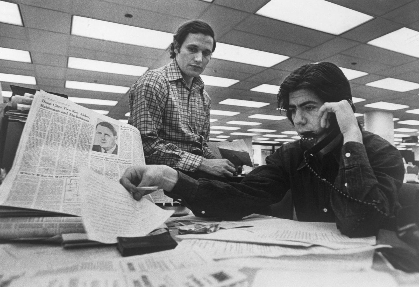 A man on the phone looking at a newspaper while his coworker stands over him.