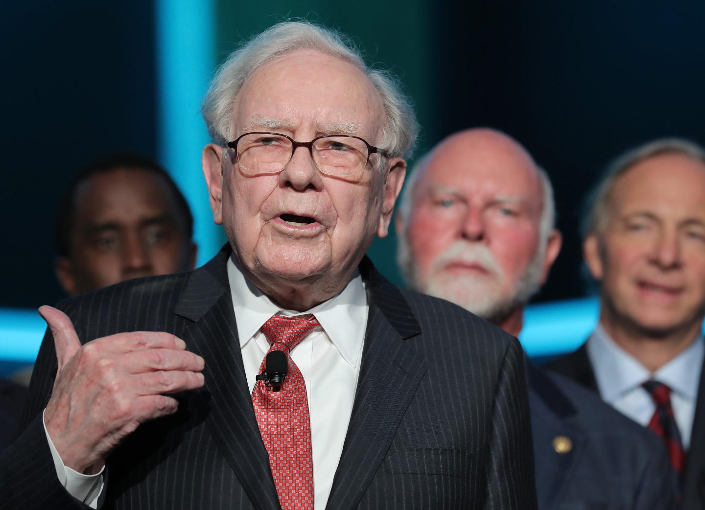 Warren Buffett gesturing with his right hand and speaking with a group behind him.