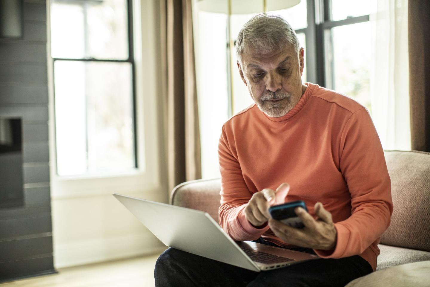 Senior man using laptop computer and smartphone at home.