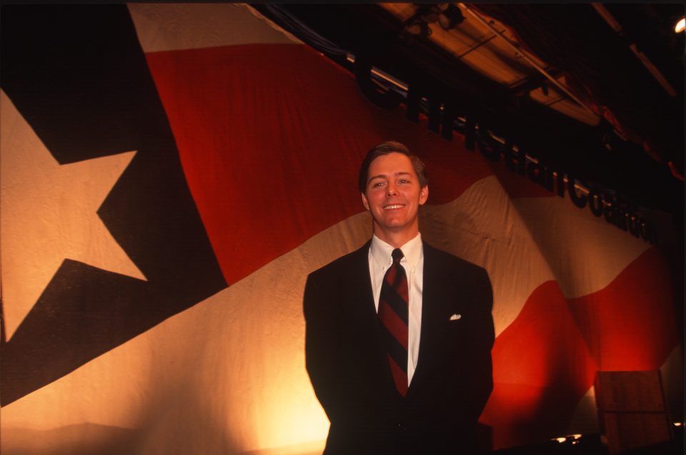 A man in a suit and tie stands in front of an American Flag.