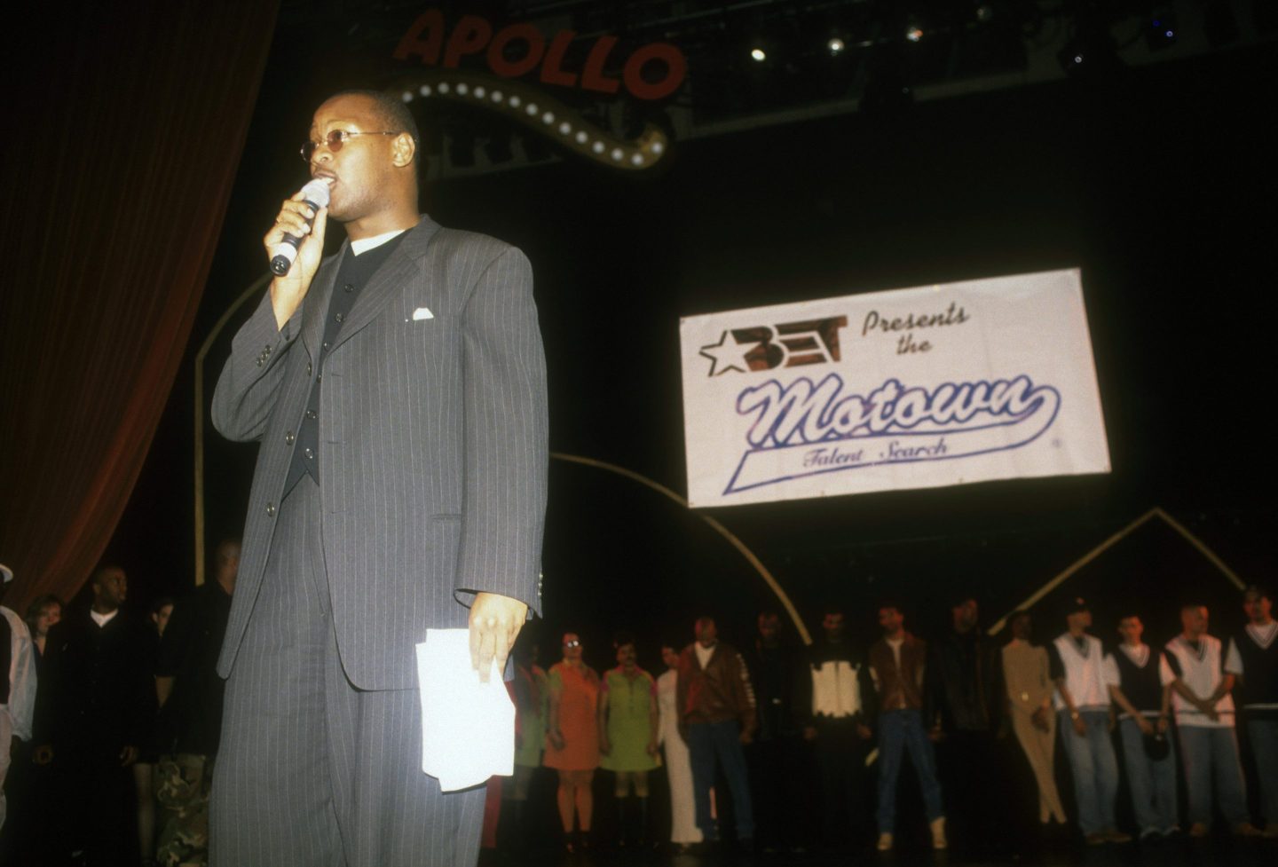 A man stands on stage with a microphone and a sign behind him reading "BET presents the Motown talent search."