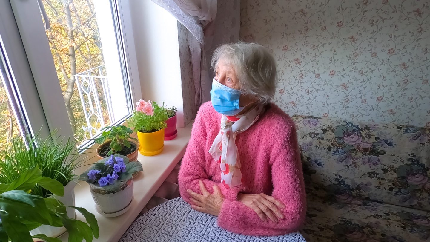 Lockdown. Covid-19, health, safety and pandemic concept &#8211; senior old lonely woman wearing protective medical mask sitting near the window at home for protection from virus