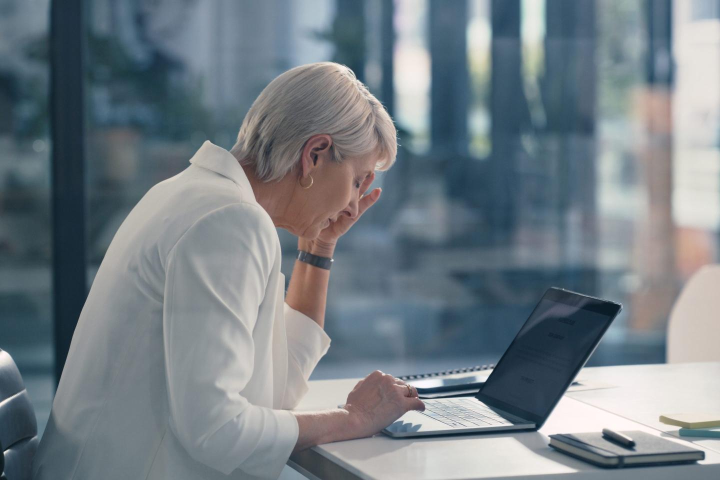 A woman seated in a chair in front of a laptop touches her temple