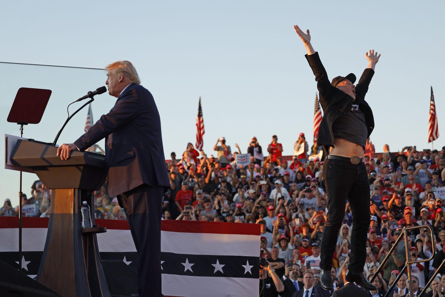 Elon Musk leaps on stage while Donald trump speaks at a podium during a campaign rally.