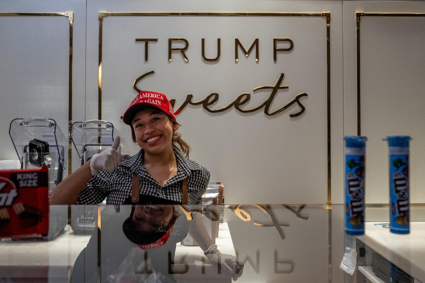 A woman wearing a MAGA hat behind a candy counter smiles and gives a thumbs up