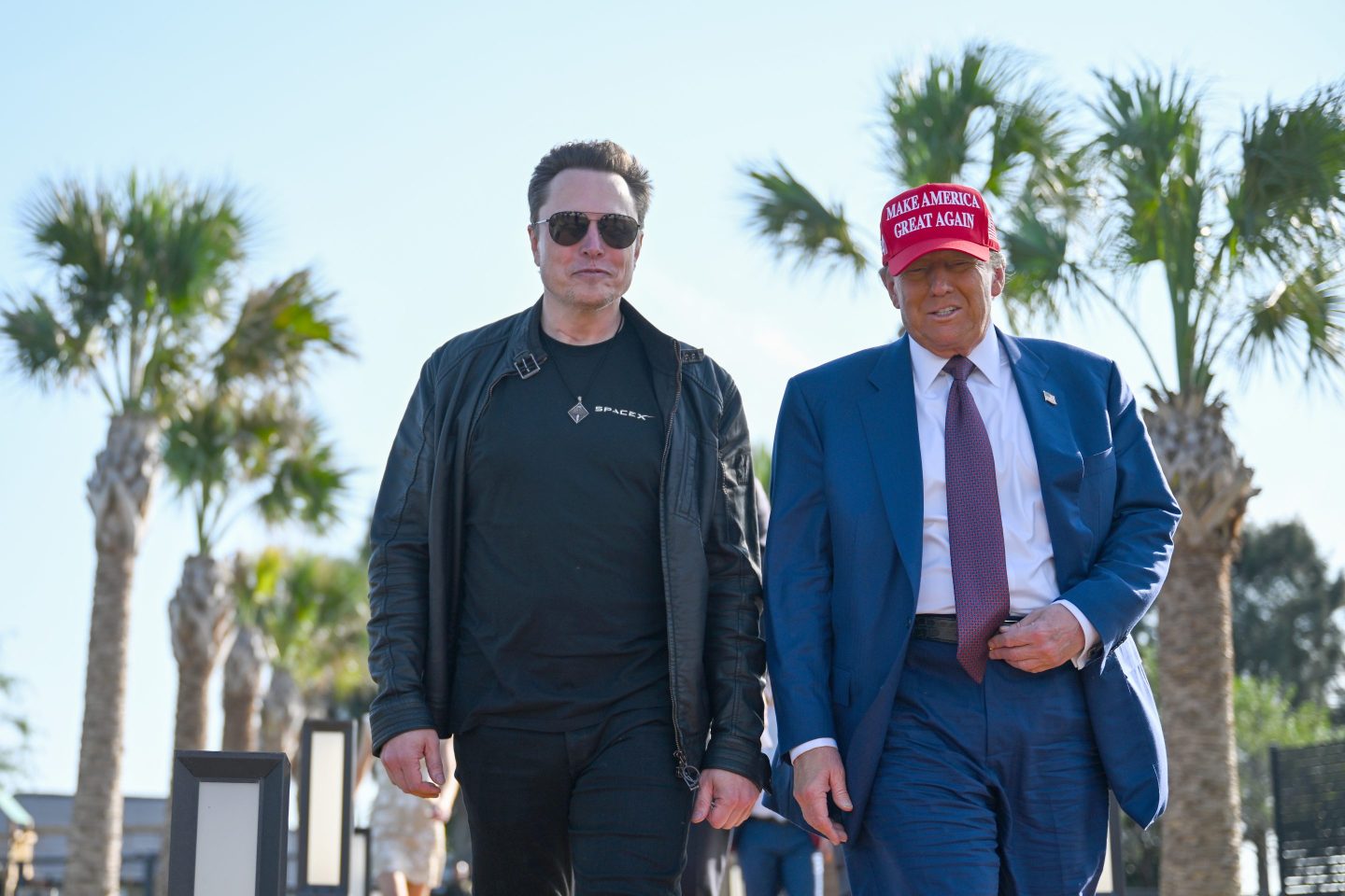 Elon Musk, dressed in black, walks with Donald Trump, dressed in a blue suit, with palm trees in the background