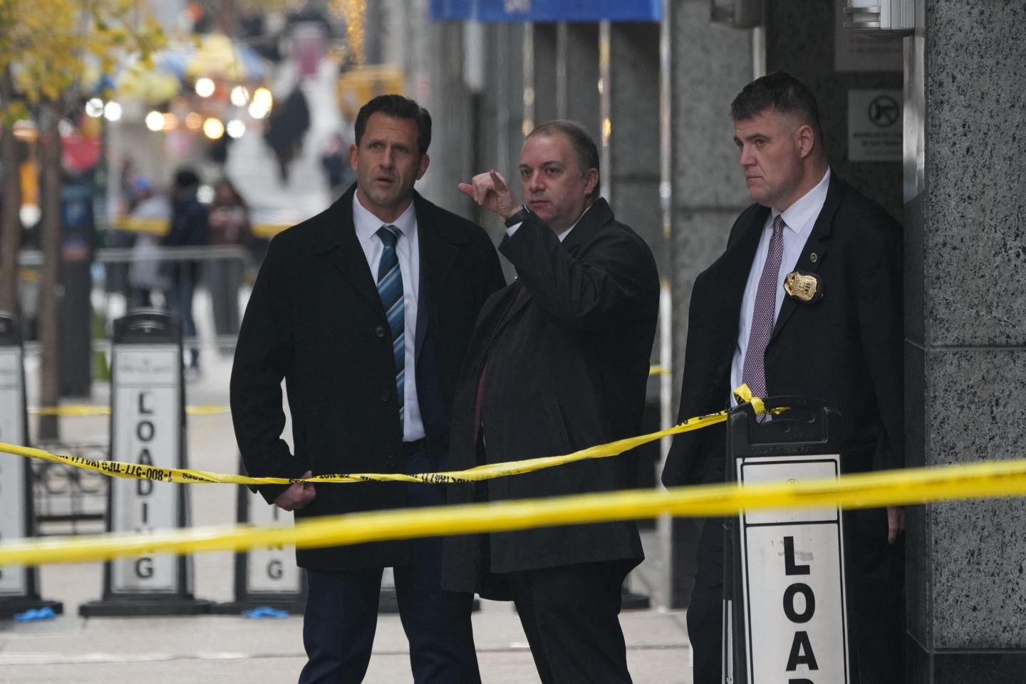 A group of investigators in suits stand behind yellow crime tape.