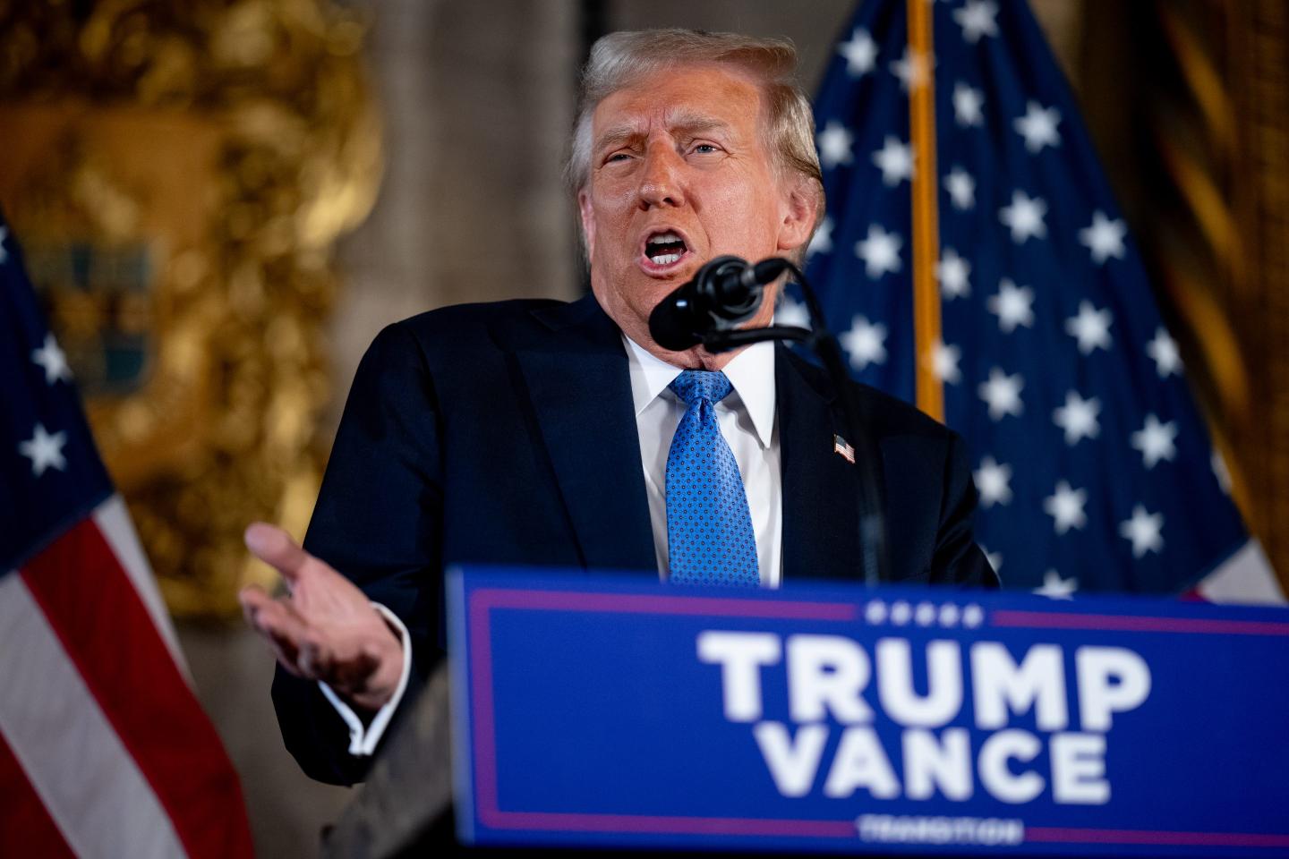 Donald Trump shrugs and gestures as he speaks into a microphone behind a "Trump Vance" podium.