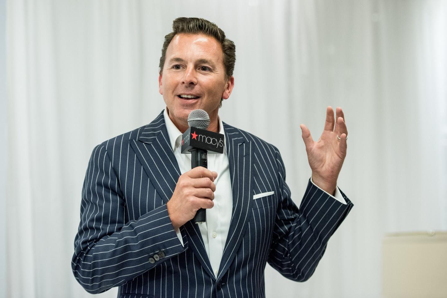 Tim Baxter is wearing a pinstripe suit and speaking into a microphone with the Macy's logo on it.