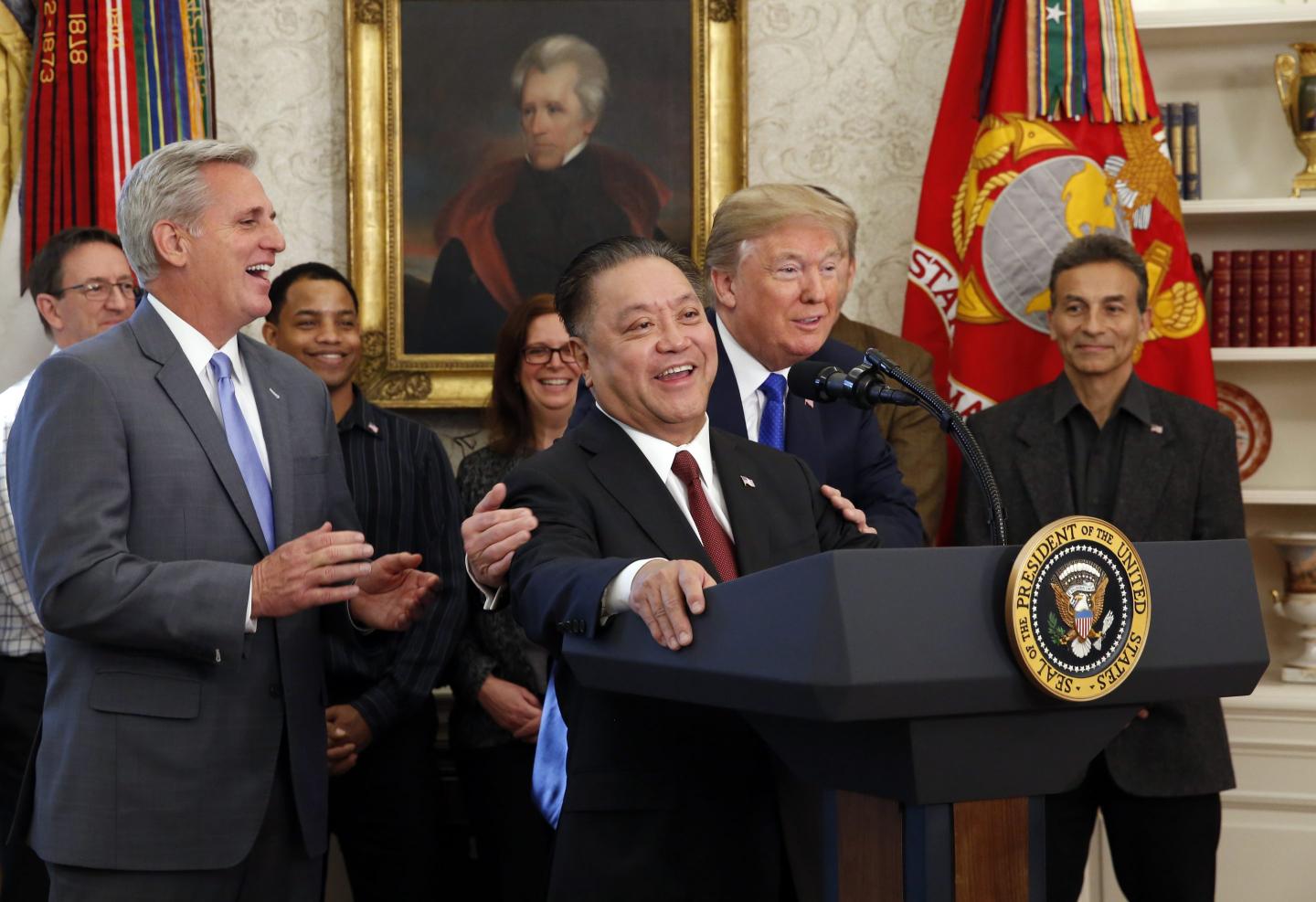 Donald Trump hugs Broadcom CEO Hock Tan from the back as Tan speaks at a podium in the Oval Office.