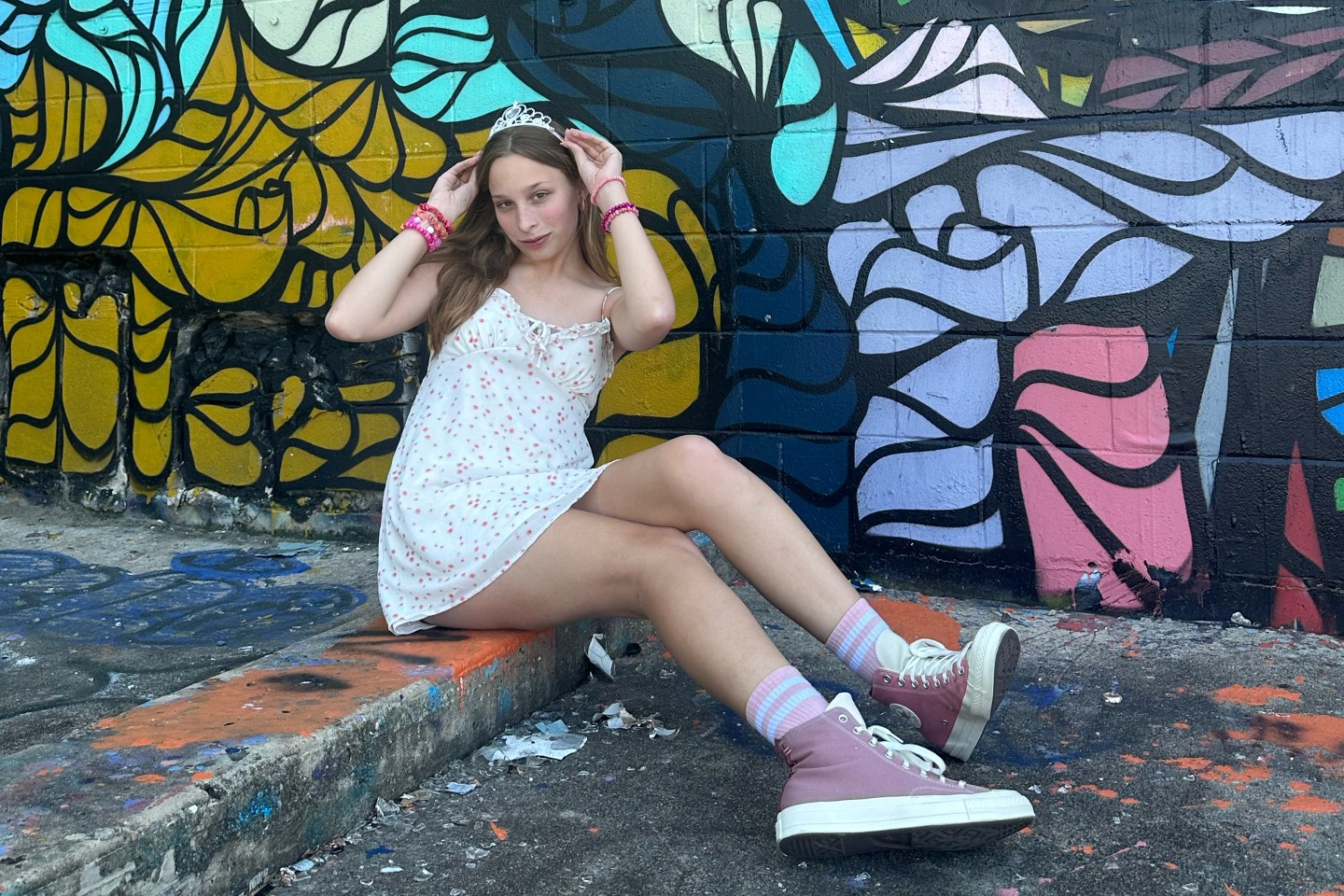 Teen girl in short dress sits on curb in front of colorful mural