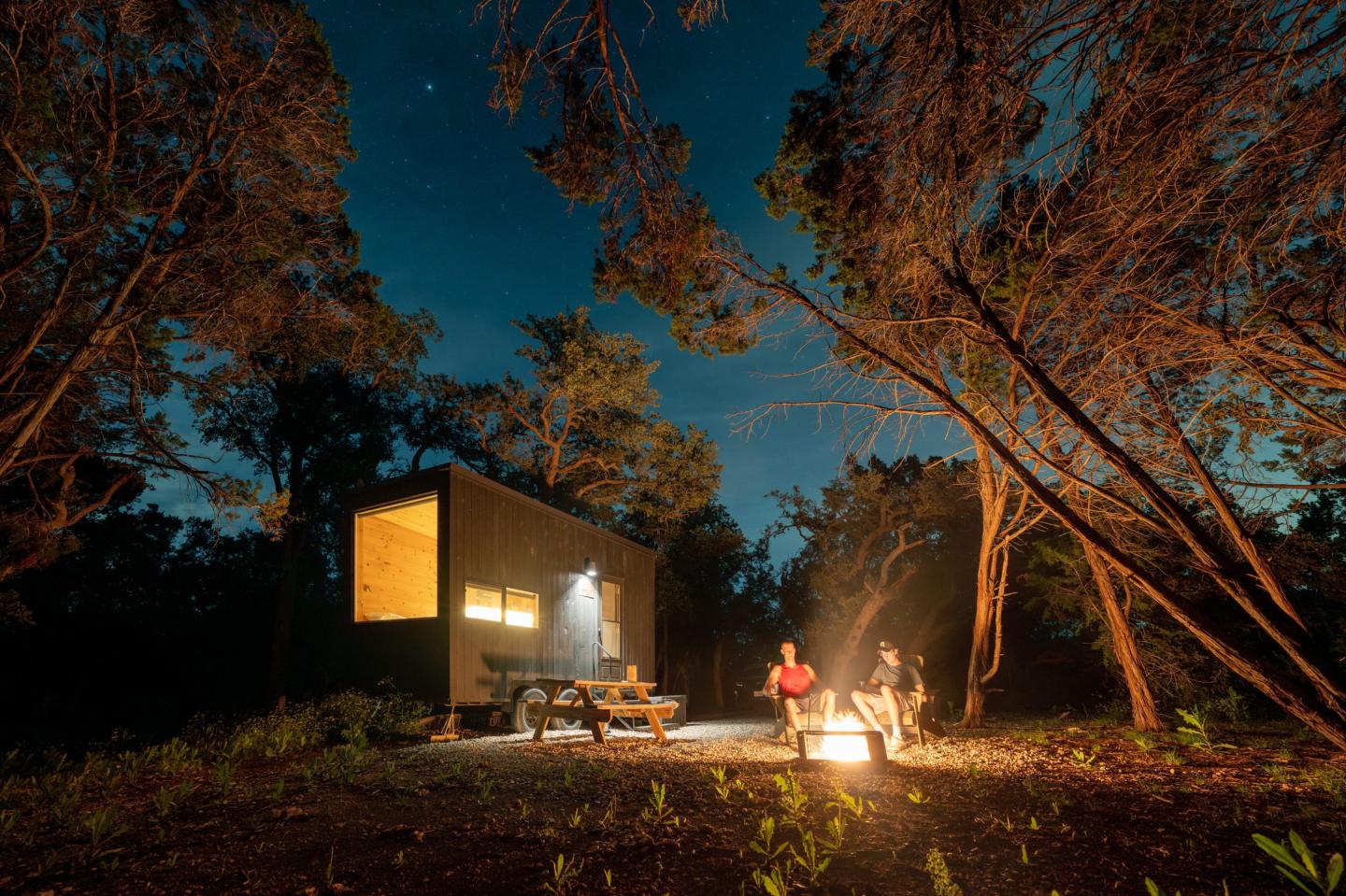 Two people sit by a fire outside a cabin