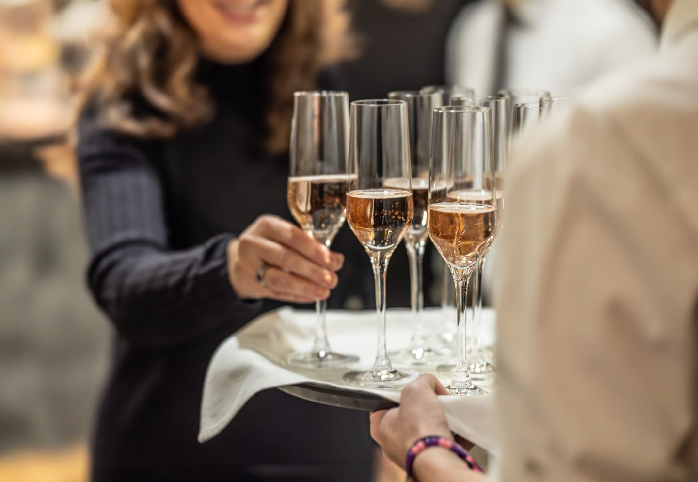 A bartender holds a plate with pink champagne and a woman in a cocktail dress takes one of the glasses at an event, party or wedding.