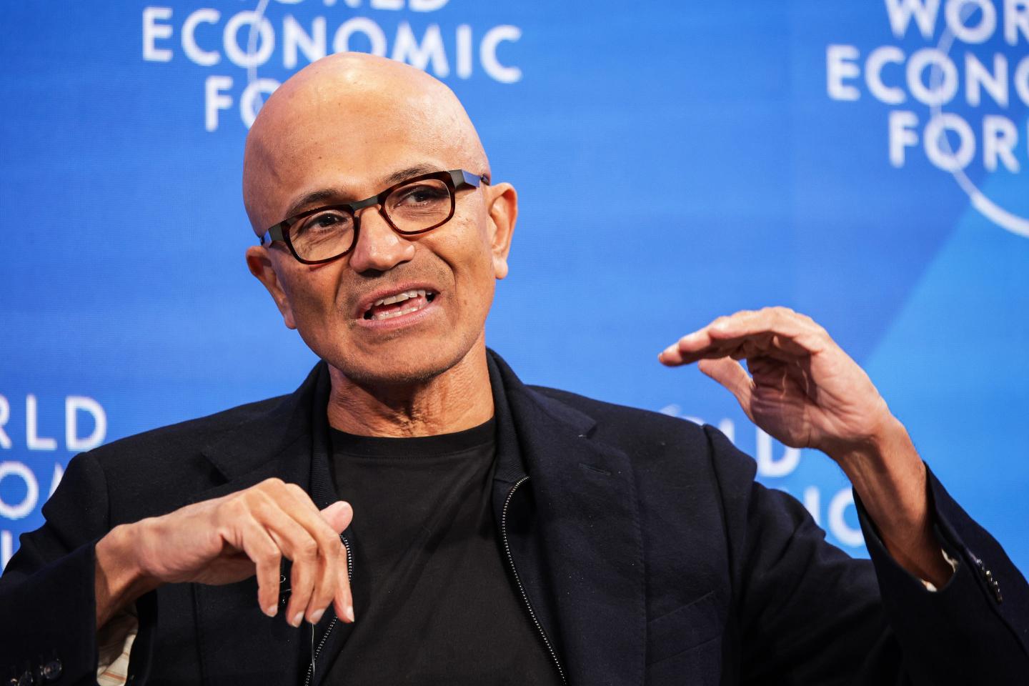 Satya Nadella gestures with his hands and speaks in front of a blue background.