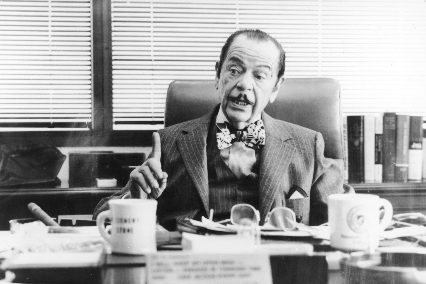 A man in a pinstripe suit and a large bowtie sits behind a cluttered desk