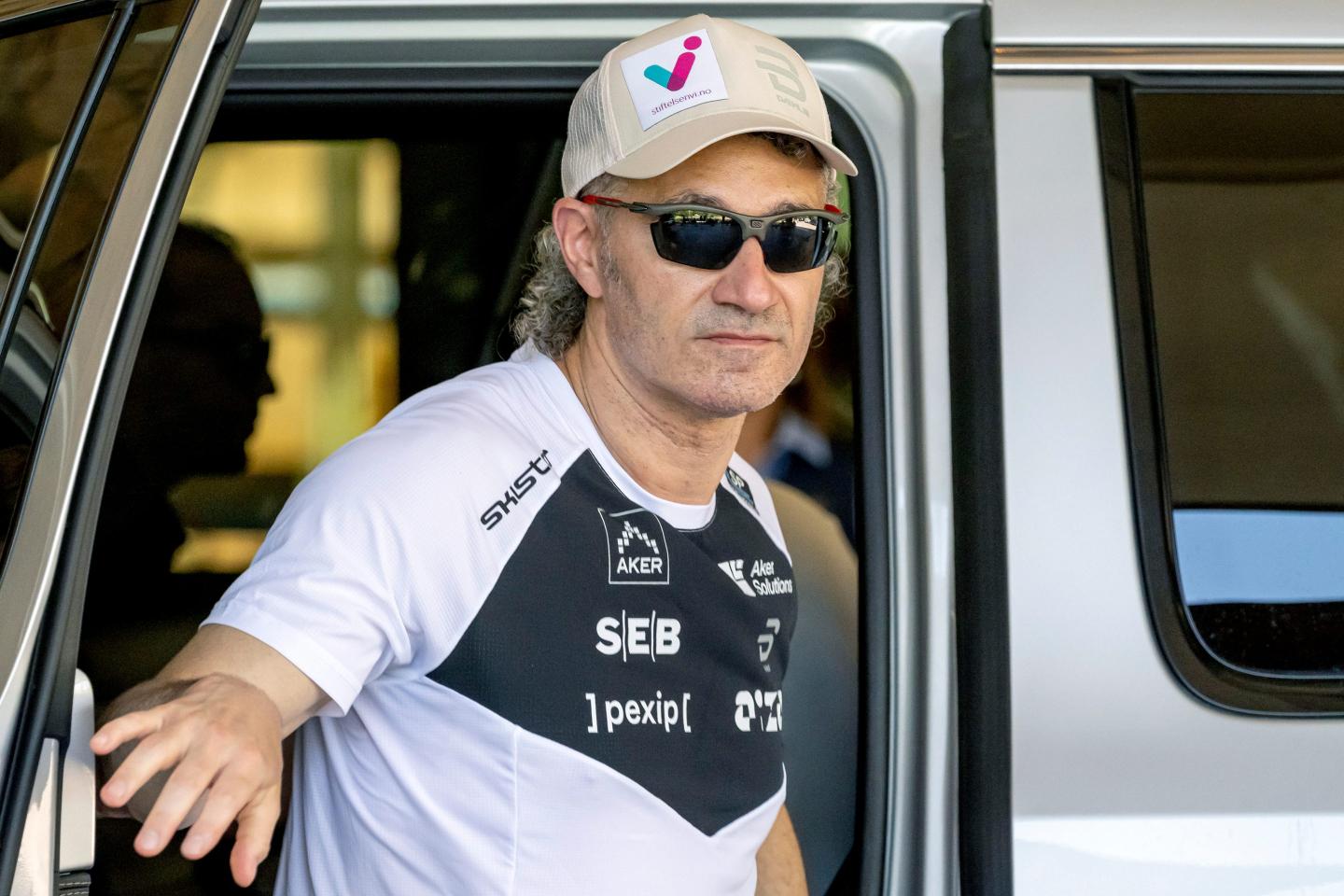 Alex Karp, wearing a t-shirt, hat, and sunglasses, opens a car door to get out.