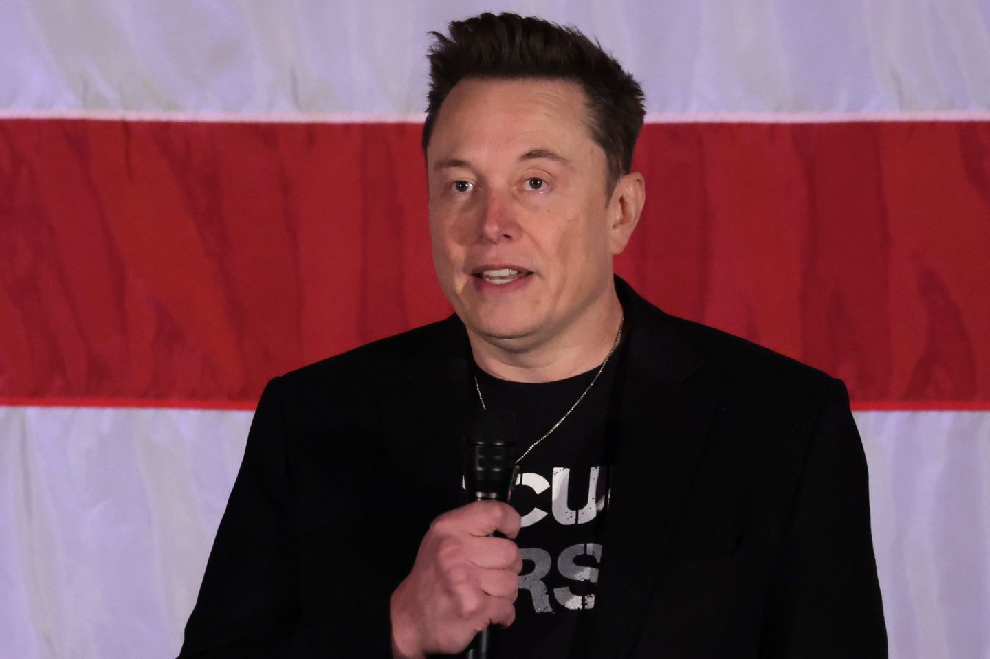 Elon Musk holds a microphone in front of the American flag