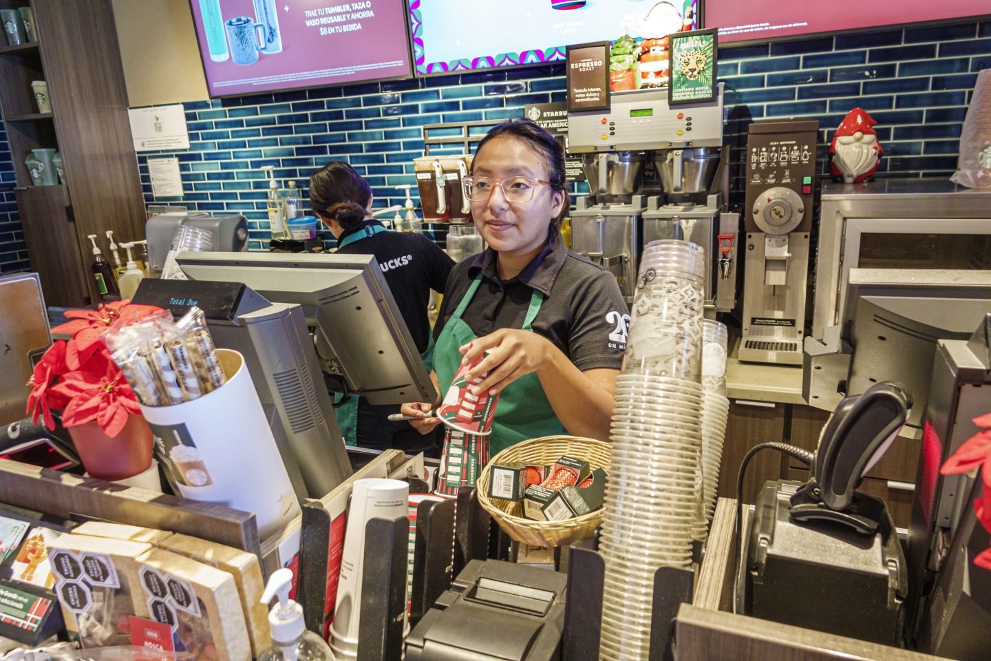 A Starbucks barista stands behind the register and grabs a drink cup from a stack.