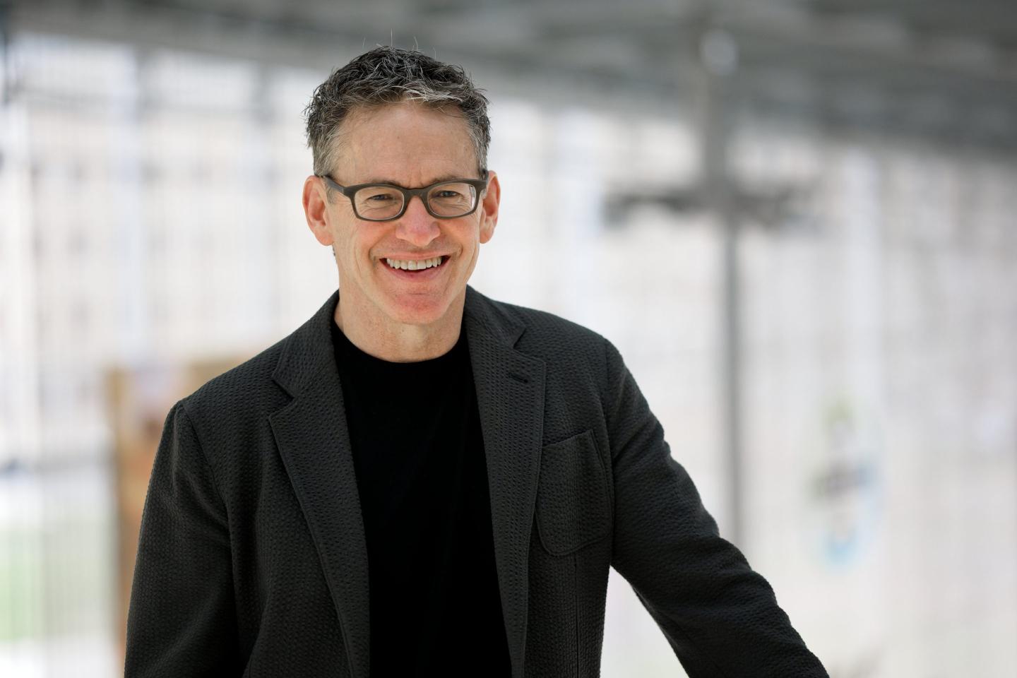 man in black shirt and black jacket, glasses poses for a portrait
