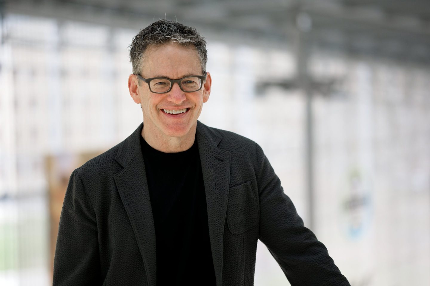 man in black shirt and black jacket, glasses poses for a portrait