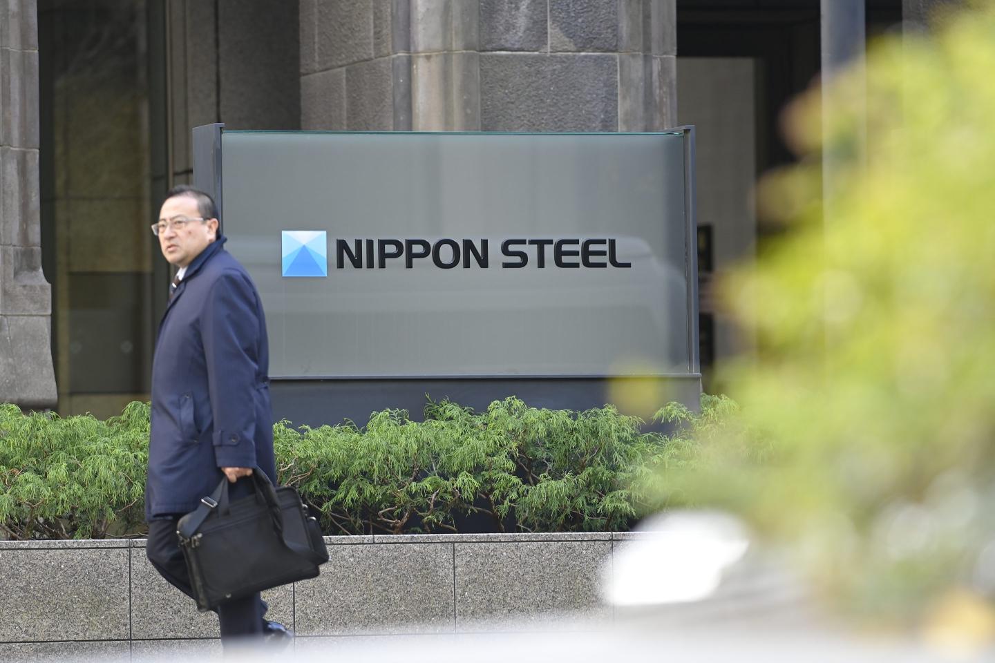 A man in a suit carrying a briefcase walks in front of a Nippon Steel sign