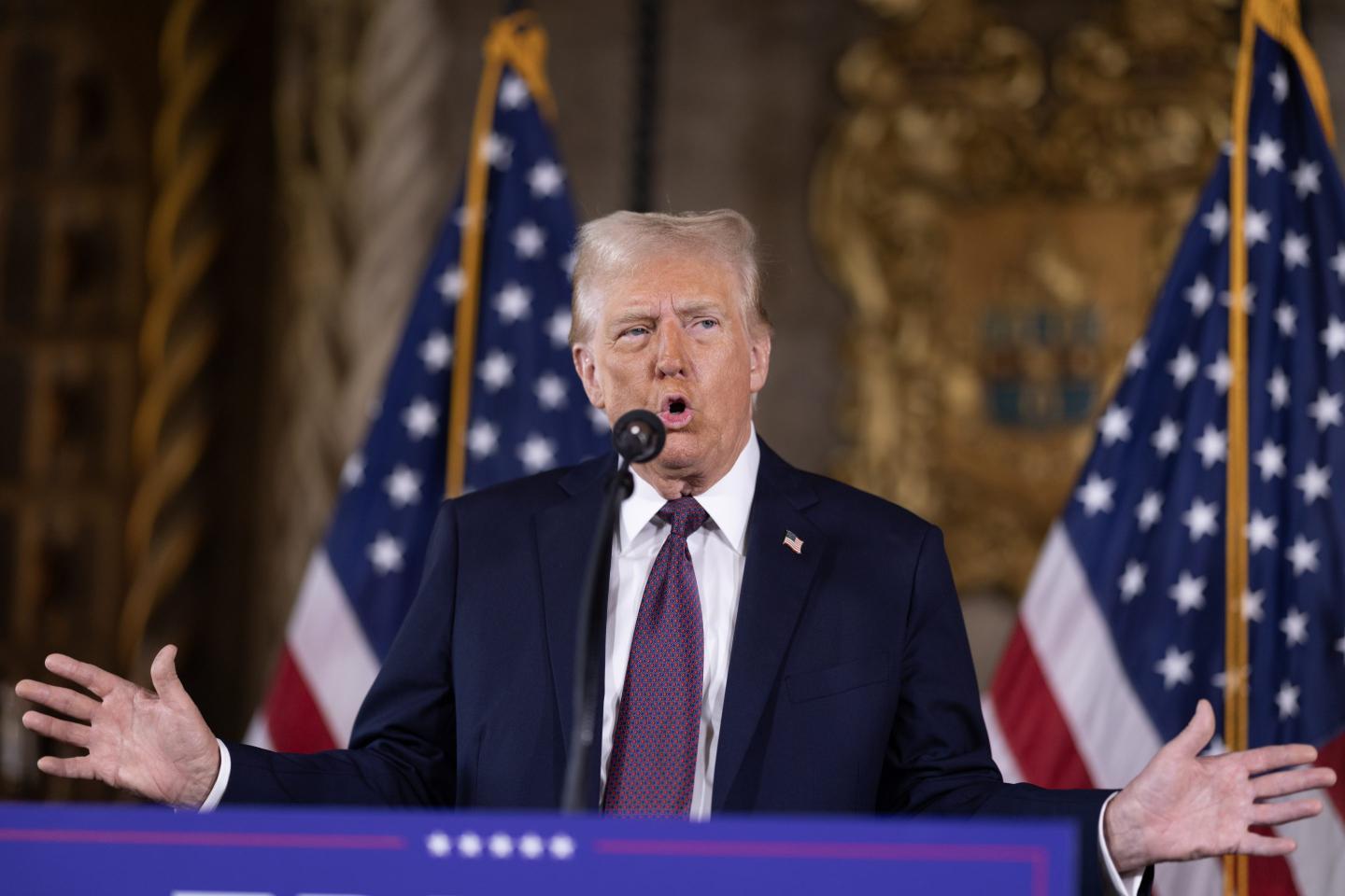 Donald Trump opens his mouth and spreads his hands as his talks at a podium with American flags in the background at a press conference.